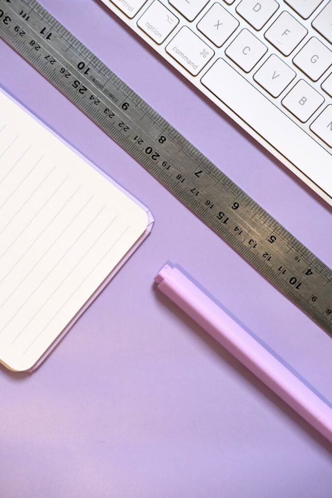 Flat lay of a minimalist workspace with keyboard, notebook, and ruler on a purple background.